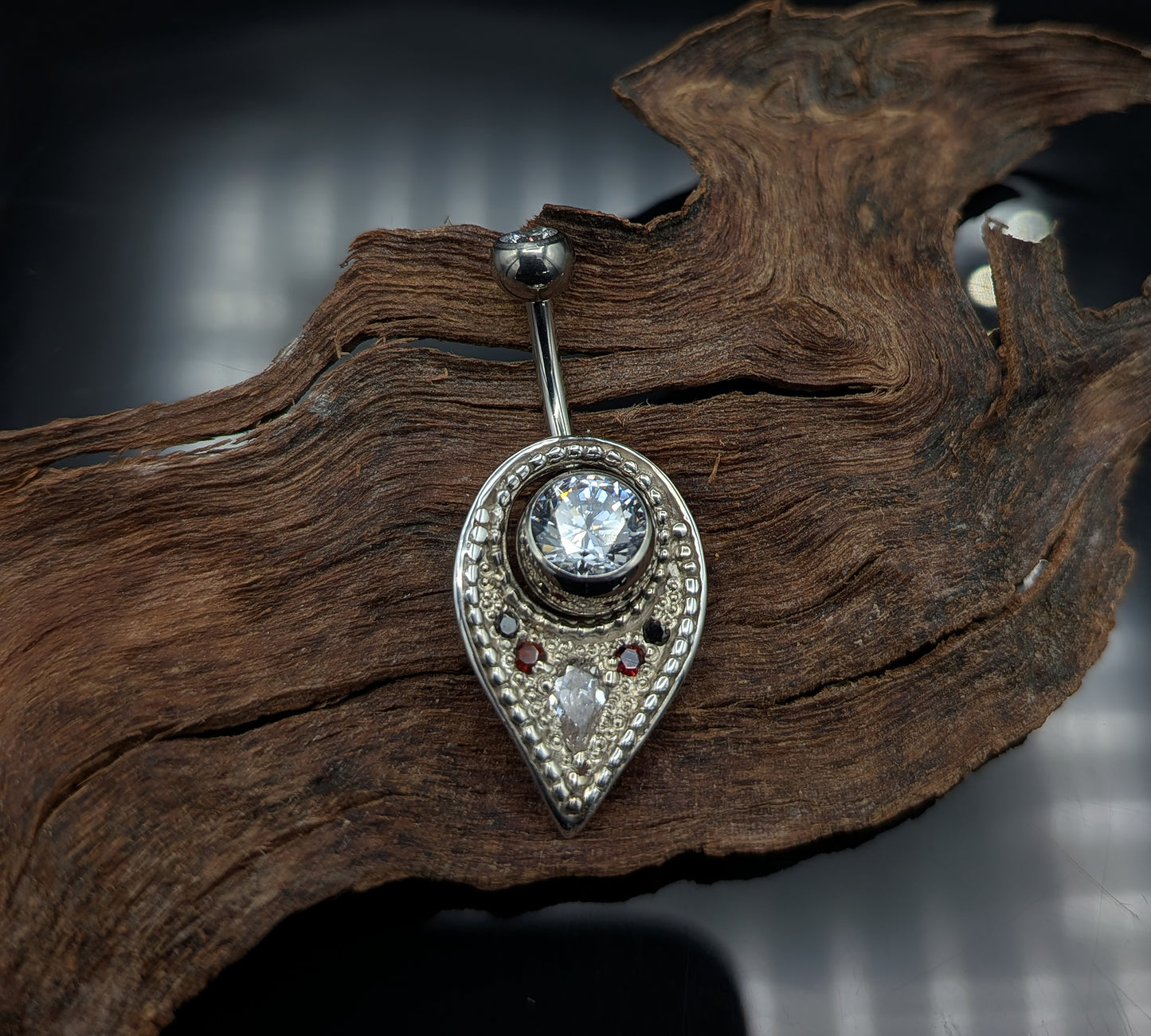 A silver navel shield decorated with granulation, set with gemstones and on a crystal navel bar