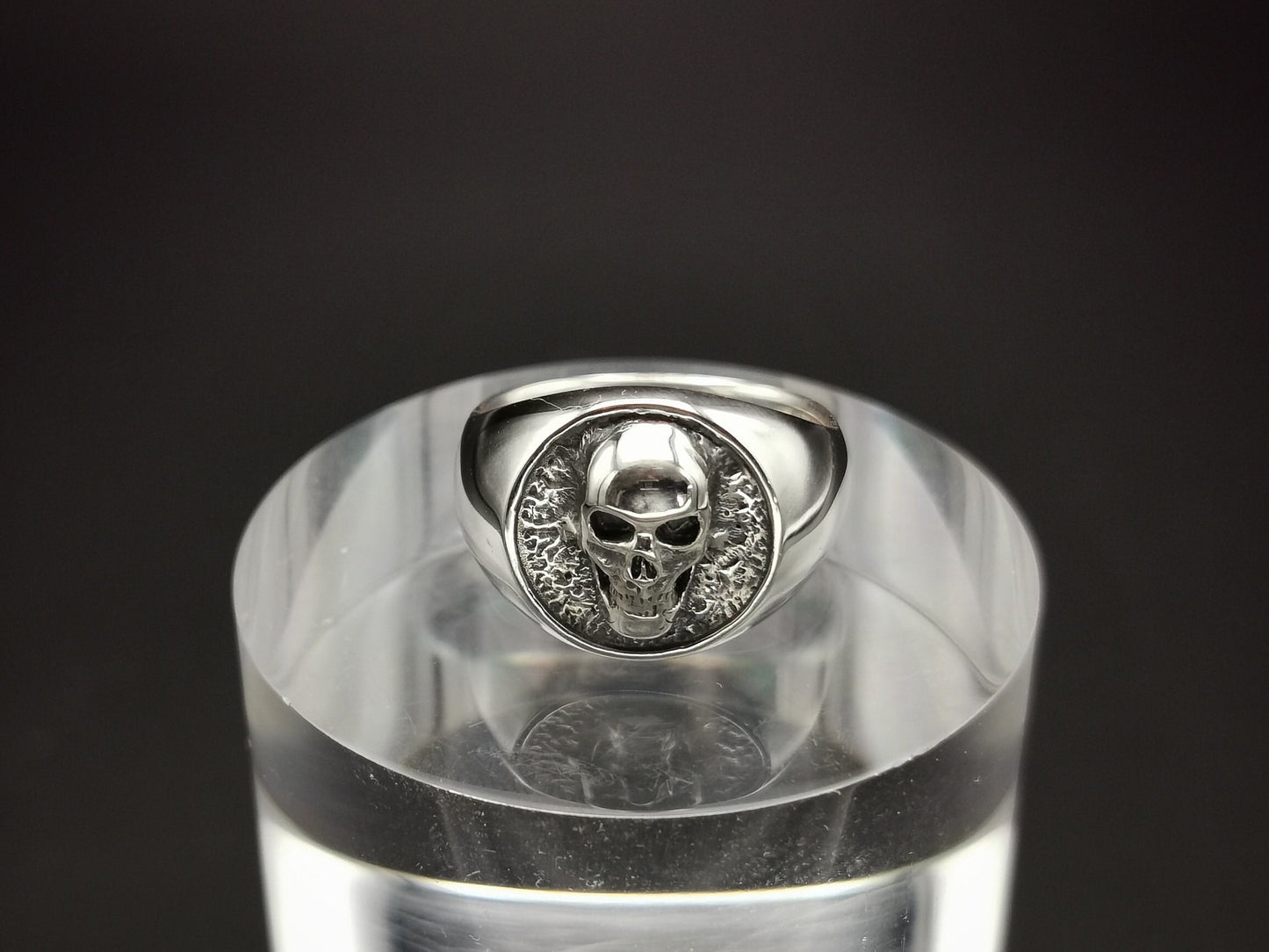 A sterling silver signet ring with a skull design, displayed on a clear stand.
