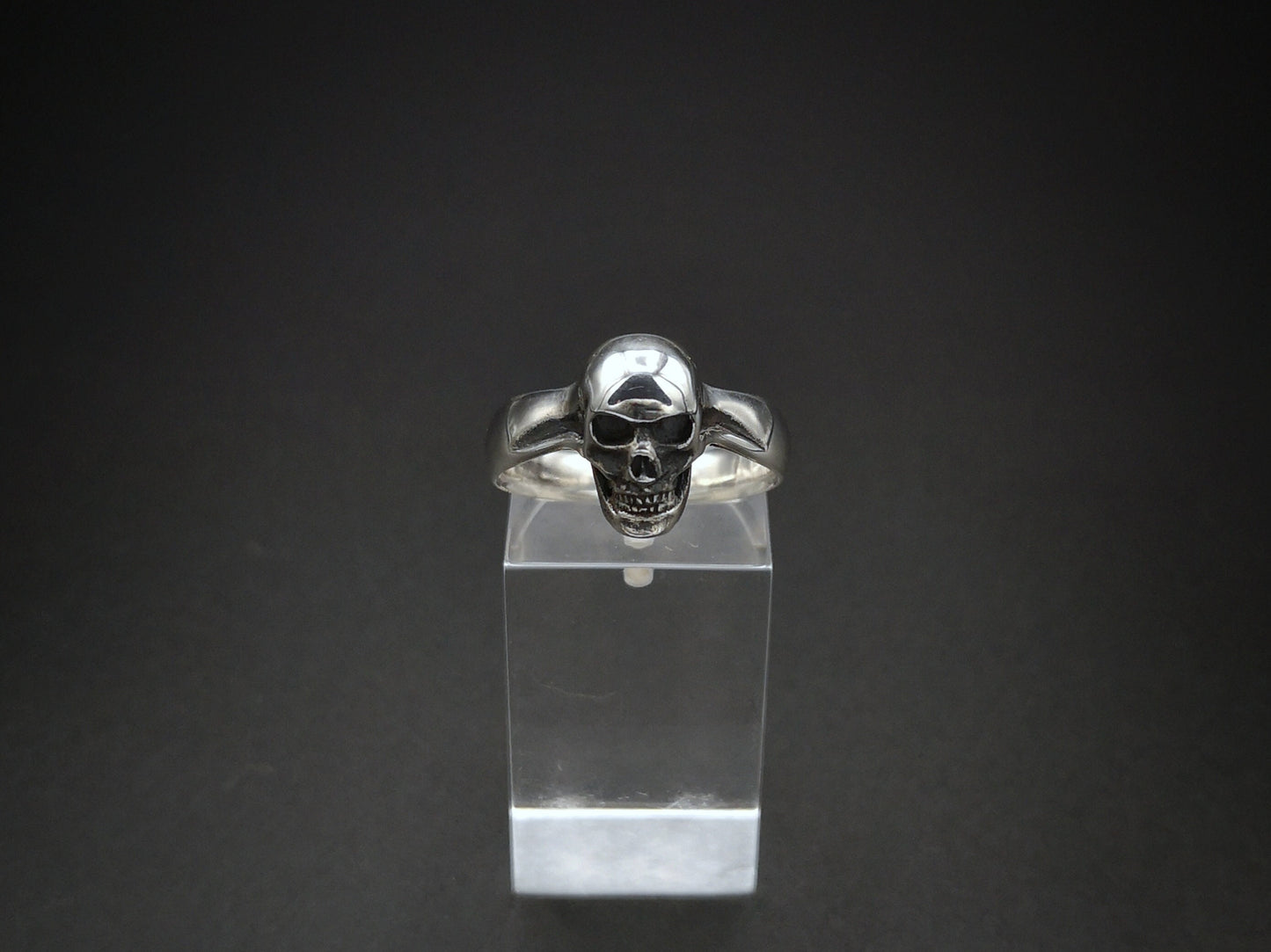 A small sterling silver skull ring displayed on a clear stand, with a detailed skull design on top.