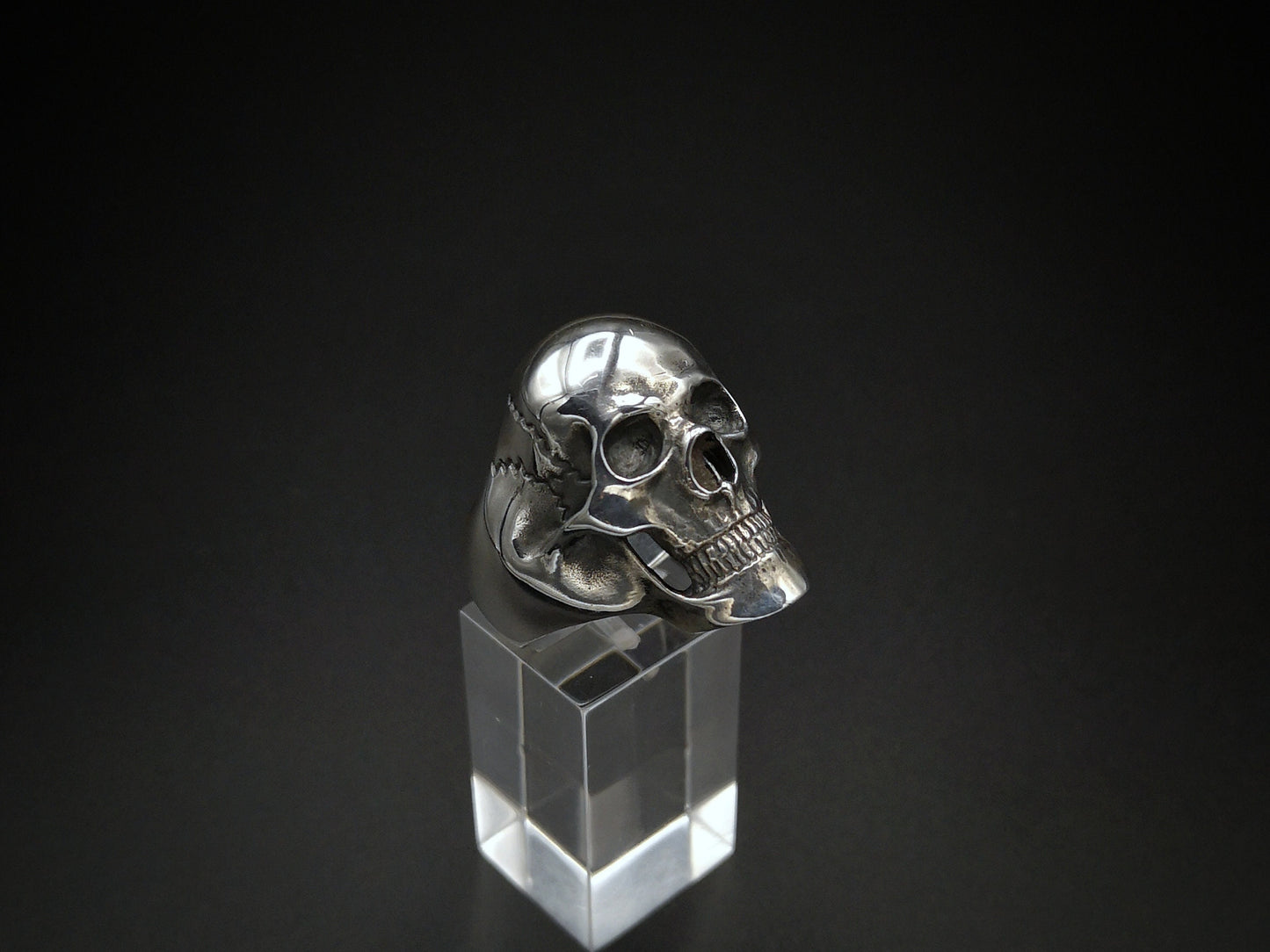 A large sterling silver skull ring displayed on a clear stand, with a polished finish.
