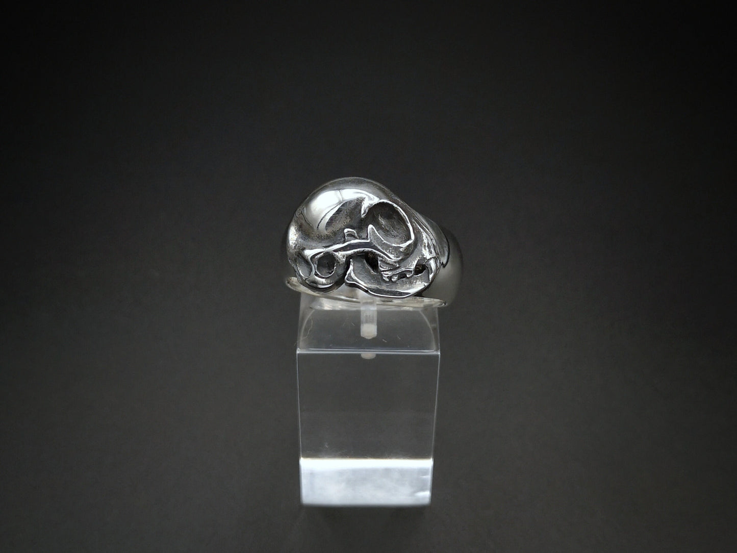 A sterling silver cat skull ring displayed on a clear stand against a dark background.
