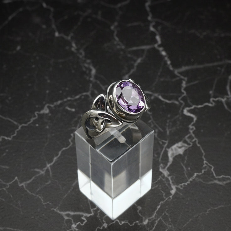 Silver ring with a purple gemstone on a clear stand against a dark marble background