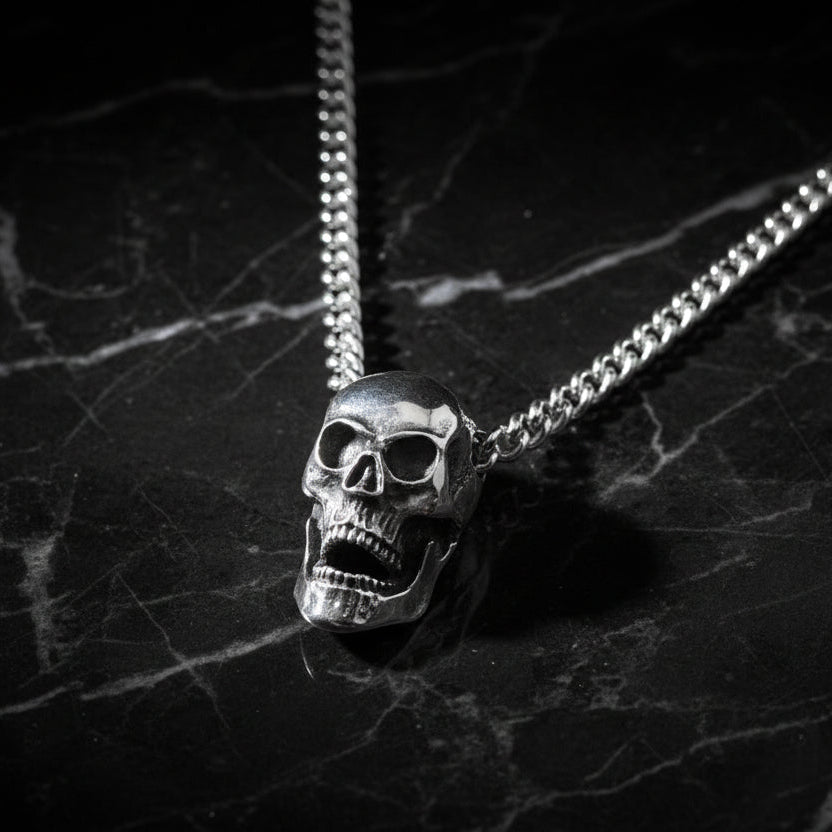 A silver skull pendant necklace with a silver curb chain, displayed on a mannequin.