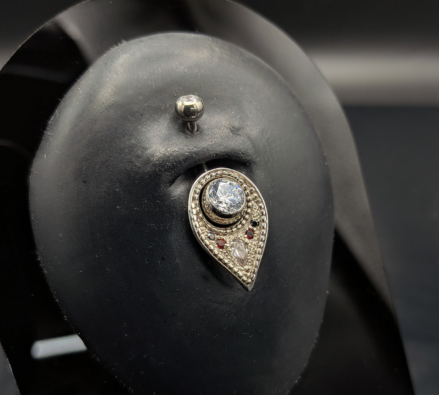 A silver navel shield decorated with granulation, set with gemstones and on a crystal navel bar