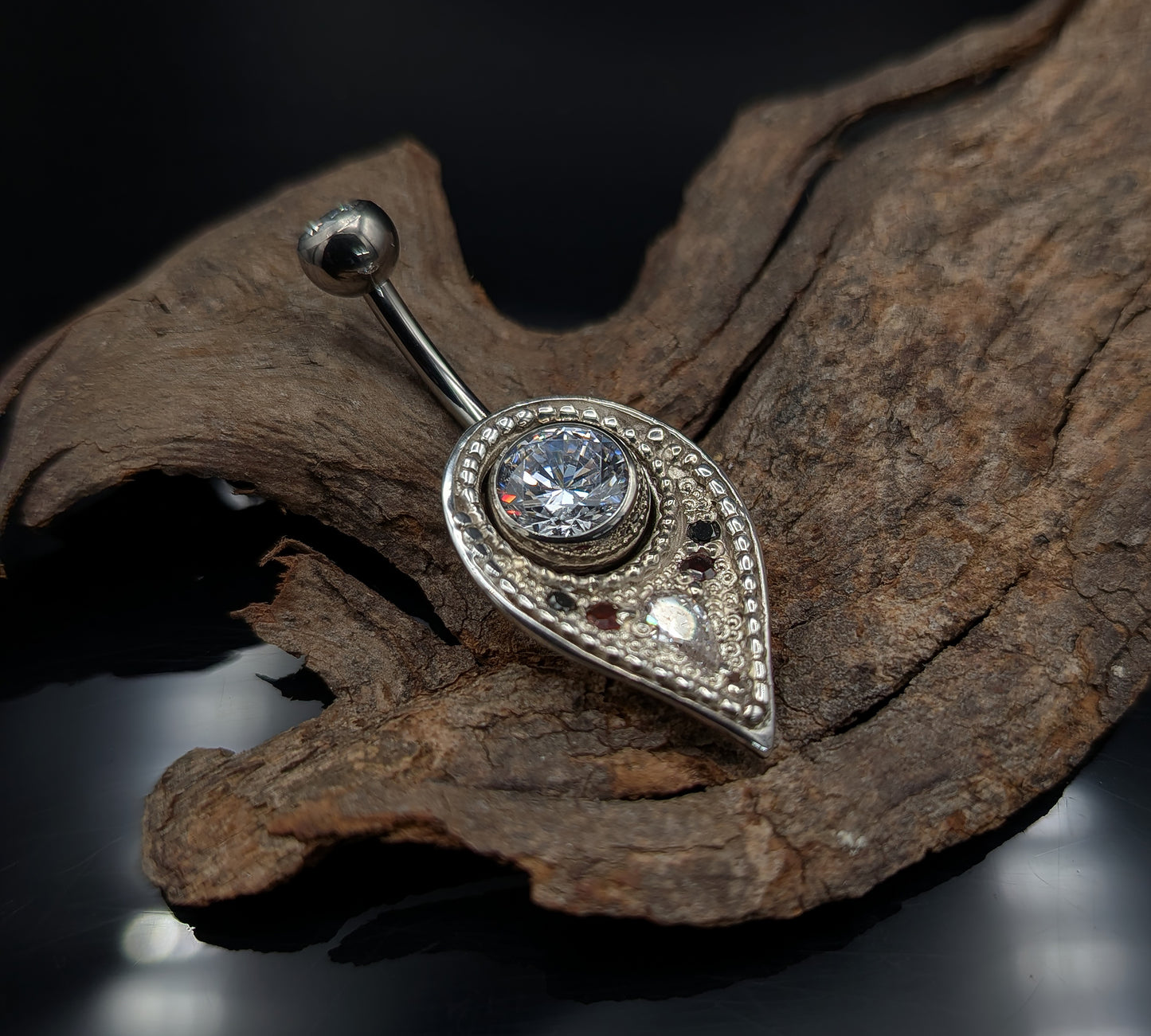 A silver navel shield decorated with granulation, set with gemstones and on a crystal navel bar