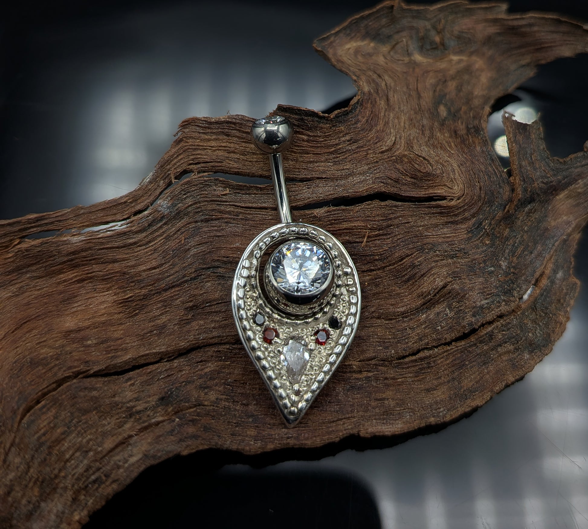 A silver navel shield decorated with granulation, set with gemstones and on a crystal navel bar
