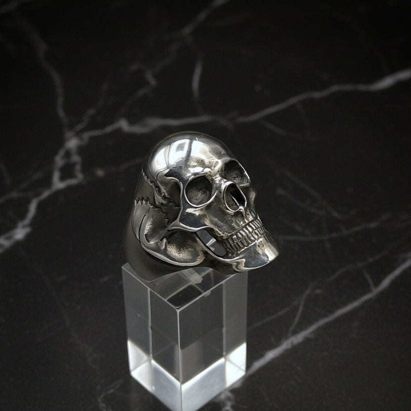 A large sterling silver skull ring displayed on a clear stand, with a polished finish.