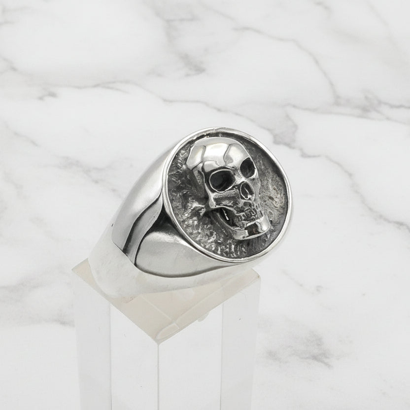 Silver skull signet ring on a clear stand against a light gray background