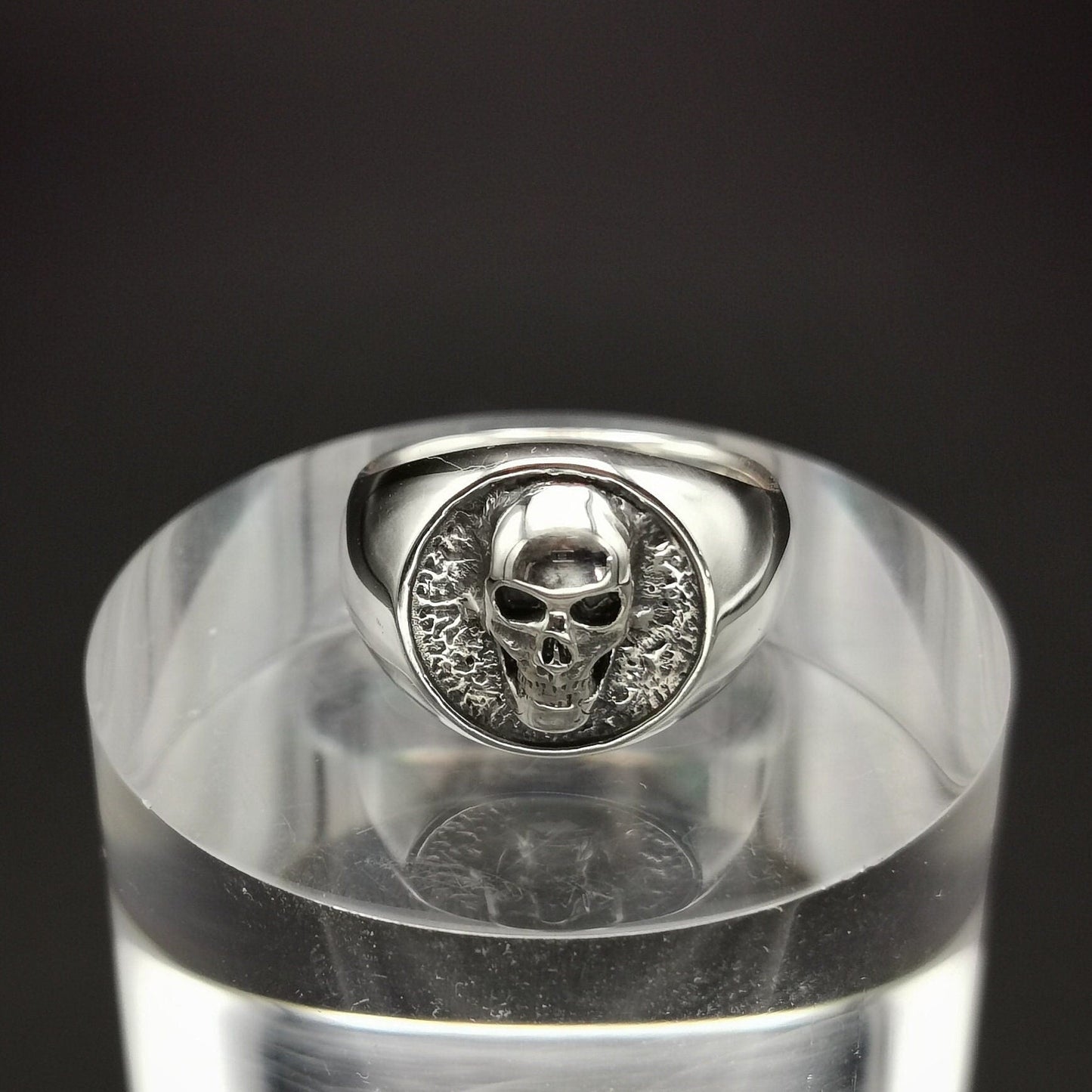 A sterling silver signet ring with a skull design, displayed on a clear stand.