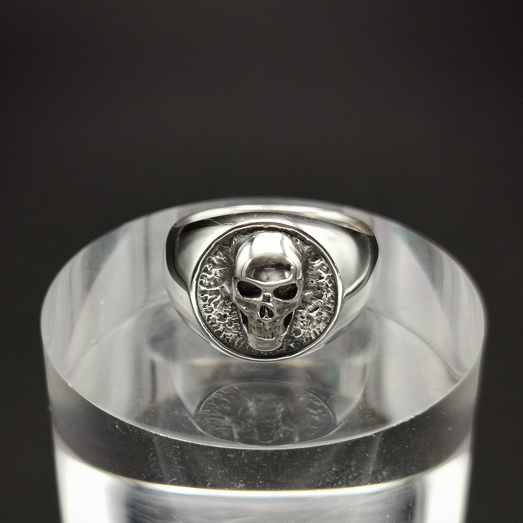 A sterling silver signet ring with a skull design, displayed on a clear stand.
