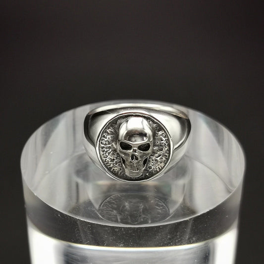 A sterling silver signet ring with a skull design, displayed on a clear stand.