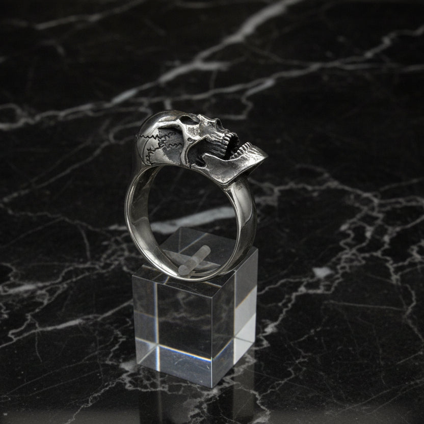 A silver screaming skull ring displayed on a clear stand, with a detailed human skull design around the finger.