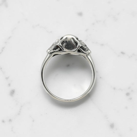 Sterling silver ring with twin skull design displayed on a clear stand against a black background.