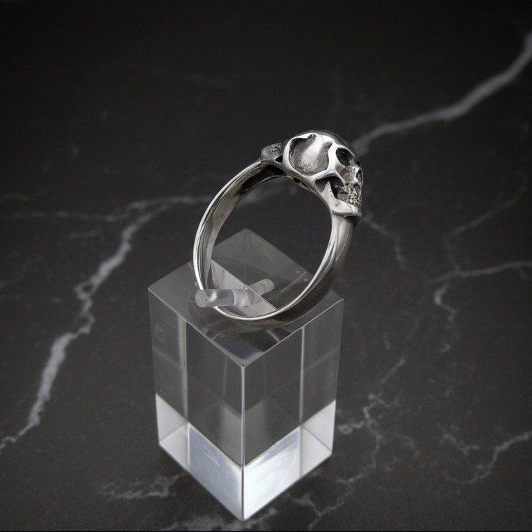 Sterling silver ring with twin skull design displayed on a clear stand against a black background.