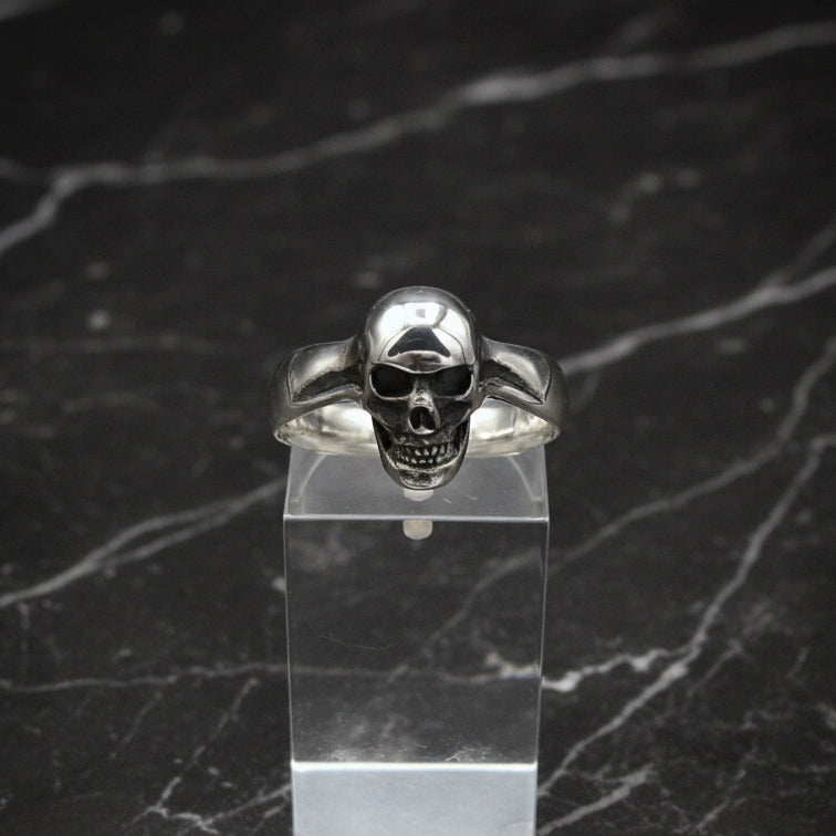 A small sterling silver skull ring displayed on a clear stand, with a detailed skull design on top.