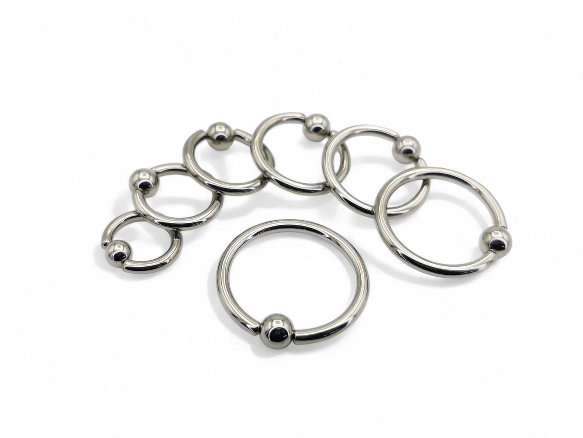 A collection of polished titanium BCRs with a ball/bead closure, displayed in two overlapping rows with varying diameters.