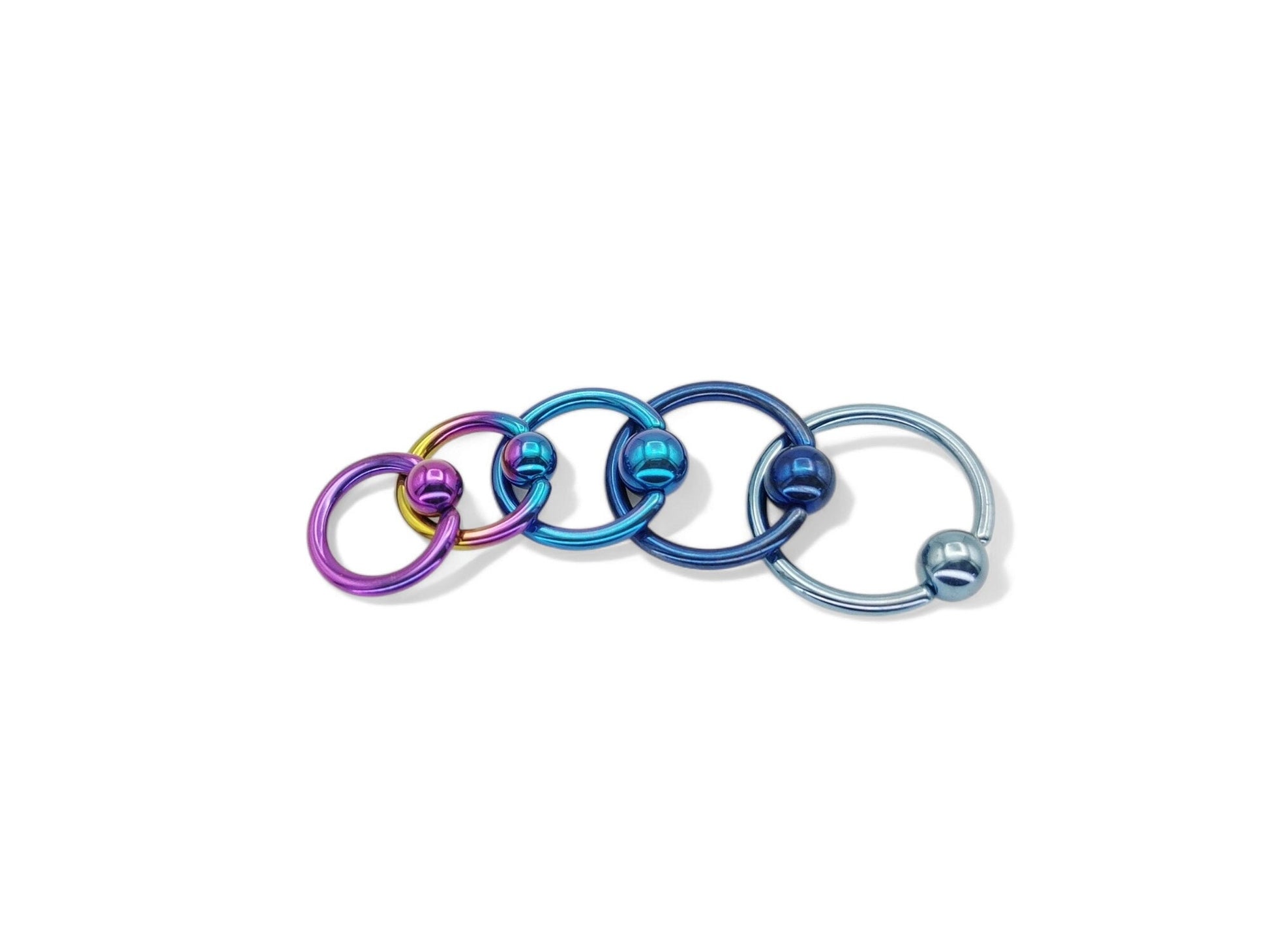A set of five titanium anodized ball closure rings in different colors: blue, turquoise, pink, and a rainbow finish, with a shiny metallic appearance.