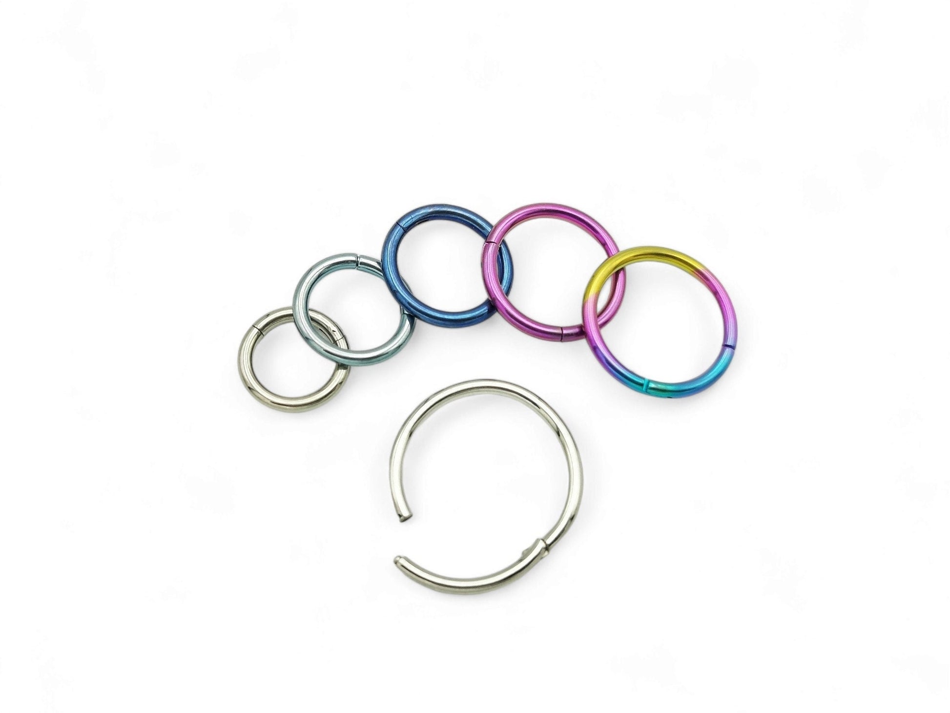 A collection of titanium clicker rings in various colors including polished silver, blue, pink, and multicolored (rainbow). They are designed for use in different types of body piercings.