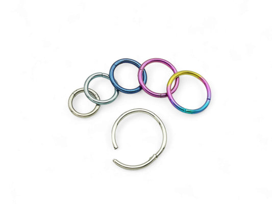 A collection of titanium clicker rings in various colors including polished silver, blue, pink, and multicolored (rainbow). They are designed for use in different types of body piercings.