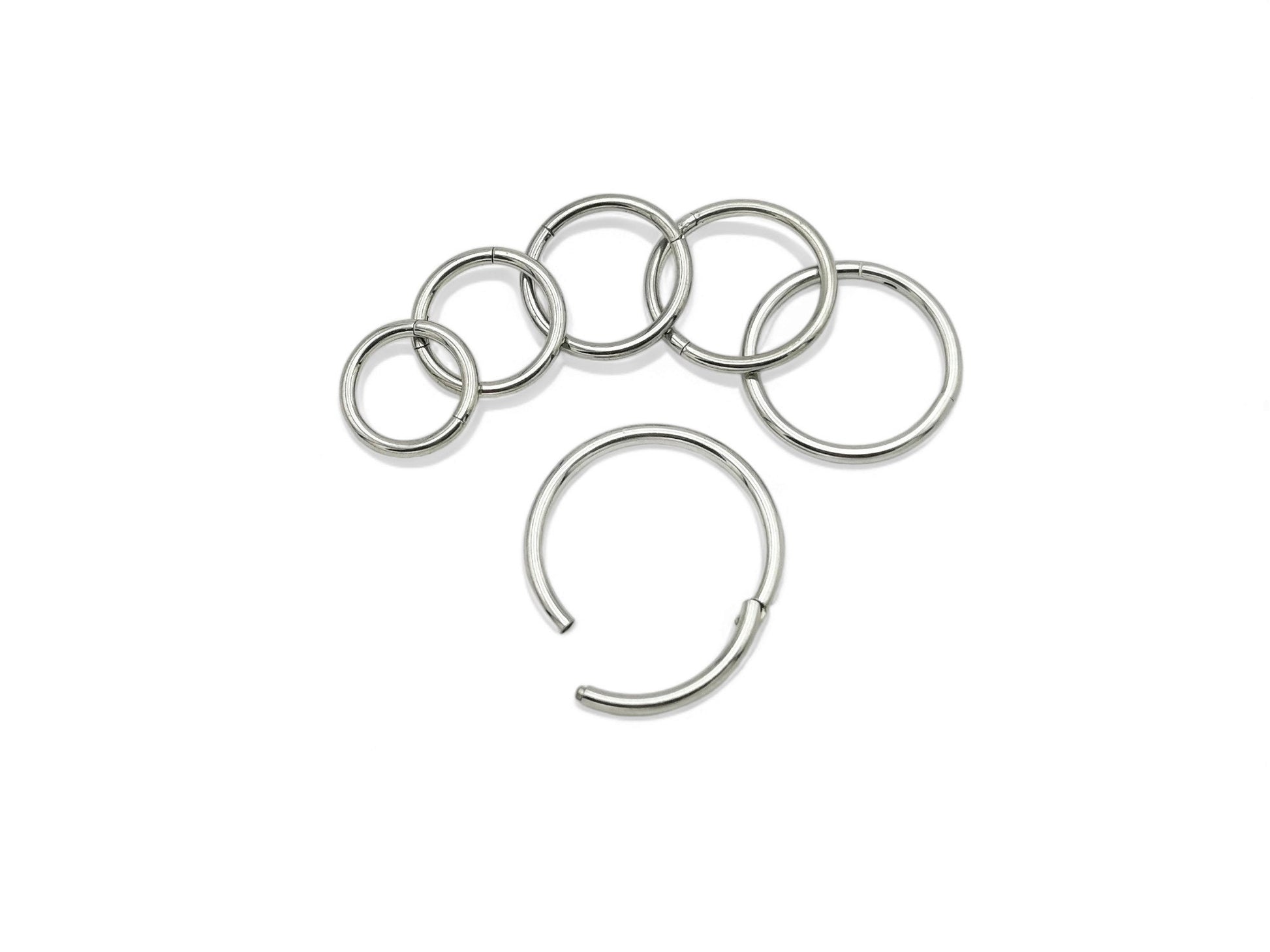 A collection of titanium clicker rings in various colors including polished silver, blue, pink, and multicolored (rainbow). They are designed for use in different types of body piercings.