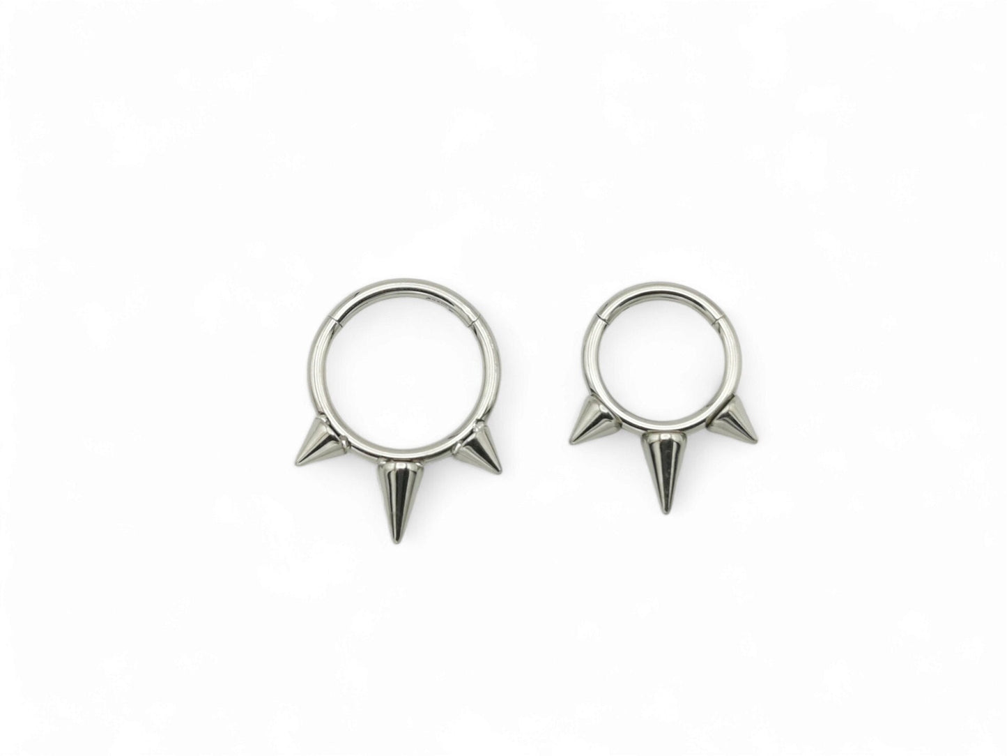 Two titanium spike clicker rings for piercings, one with a longer spike and one with a shorter spike, both with a shiny metallic finish.