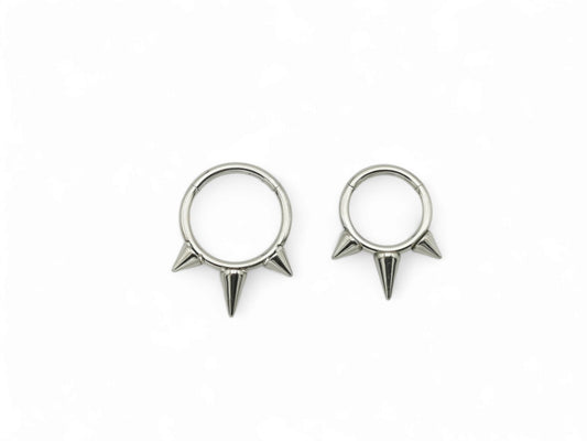 Two titanium spike clicker rings for piercings, one with a longer spike and one with a shorter spike, both with a shiny metallic finish.